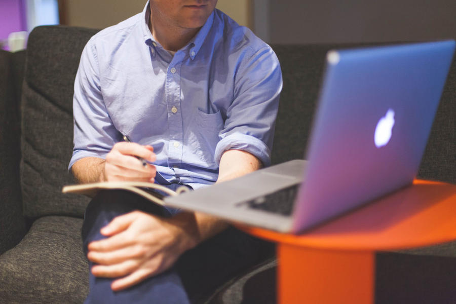 A person in a blue shirt takes handwritten notes while looking at a Macbook laptop